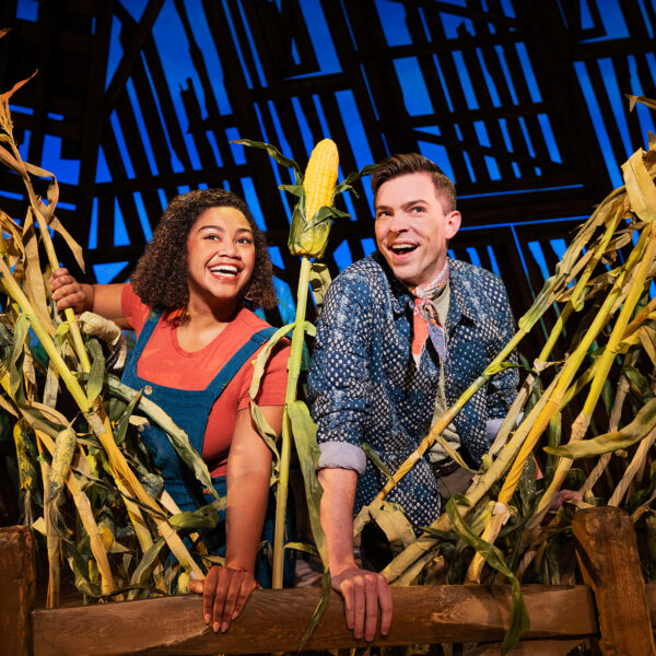 Twee performers glimlachen en leunen over een houten hek omringd door hoge maïsstengels op een podium met een schuurthema uit de musical Shucked.
