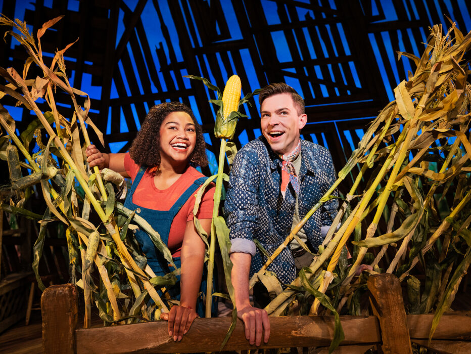 Two performers smile and lean over a wooden fence surrounded by tall cornstalks on a barn-themed stage set from the musical Shucked.