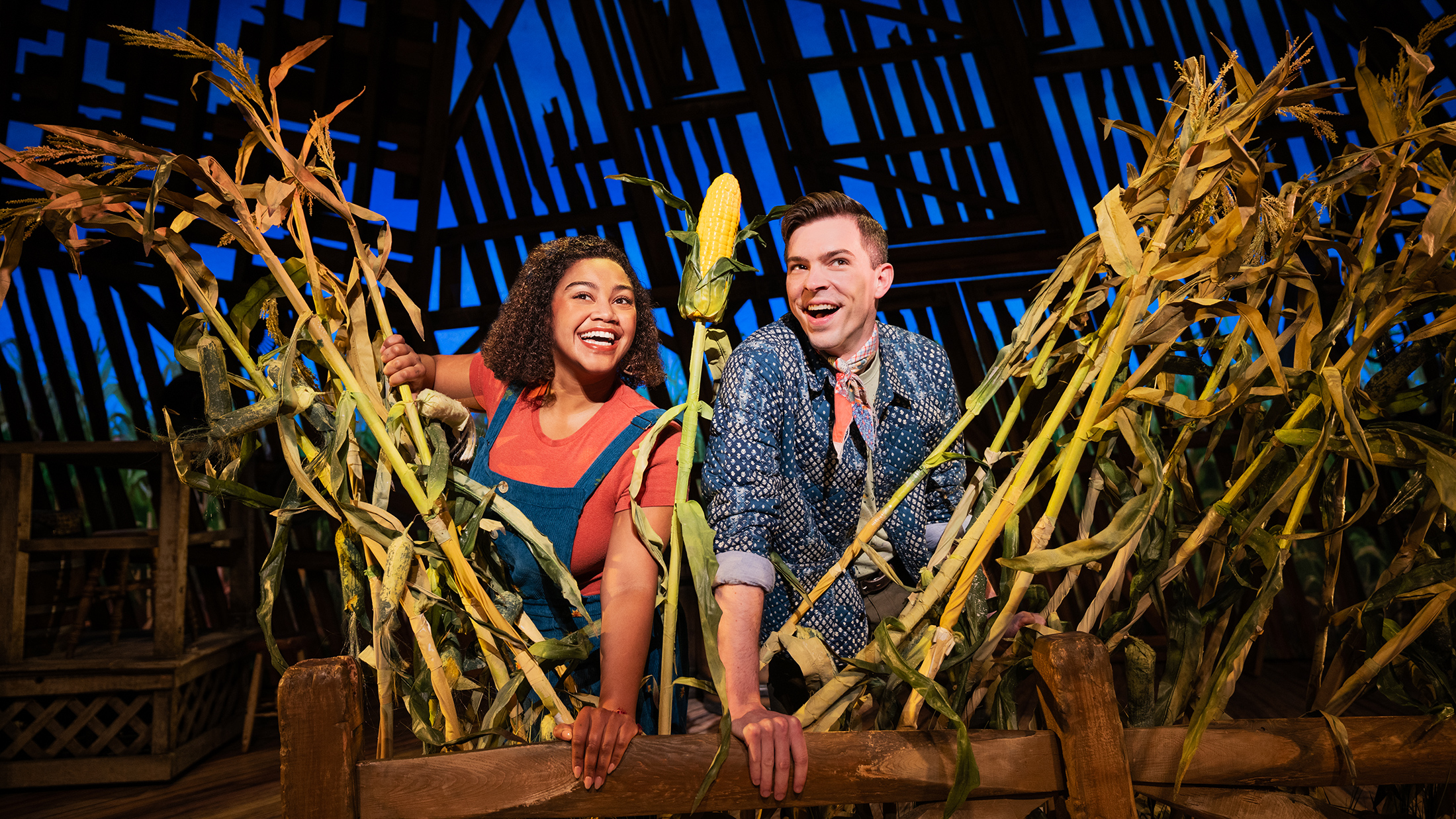 Dos artistas sonríen y se asoman por encima de una cerca de madera rodeada de altas mazorcas de maíz en un escenario ambientado en un granero del musical Shucked.