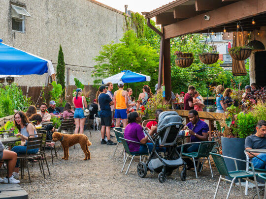 La gente disfruta de bebidas y aperitivos en un exuberante jardín en el PHS Pop Up Garden de South Street. El jardín cuenta con mesas a la sombra y macetas colgantes.