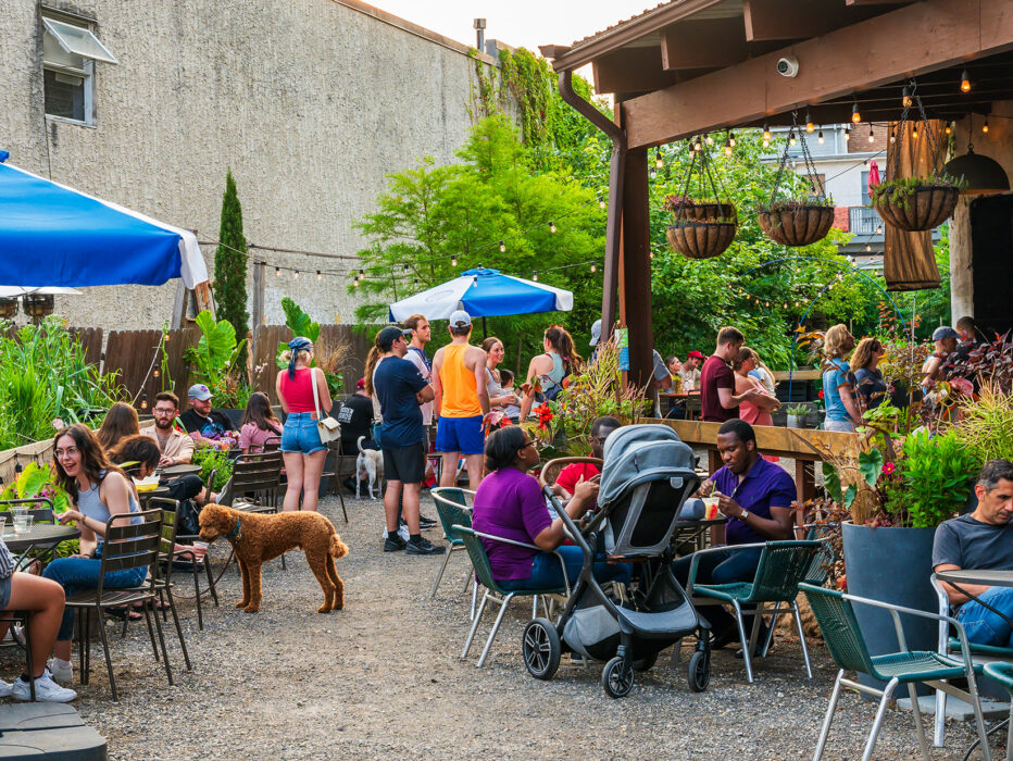People enjoy drinks and bites in a lush garden setting at the PHS Pop Up Garden on South Street. The garden is complete with shaded tables and hanging planters.