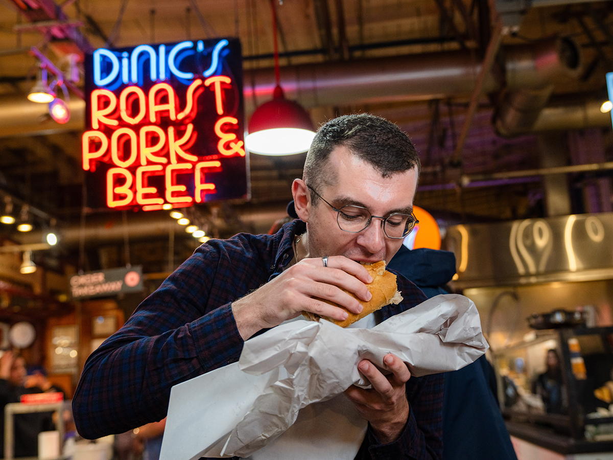 A man takes a bite of a roast pork sandwich wrapped in paper at DiNic’s, with a glowing neon “Roast Pork & Beef” sign behind him.