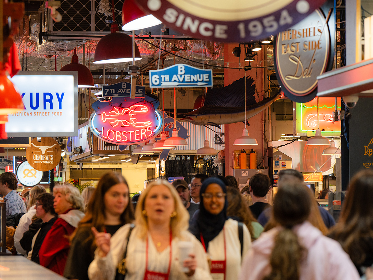 Shoppers fill the aisles of Reading Terminal Market beneath hanging neon signs and vendor placards, including a bright “Lobsters” sign overhead.