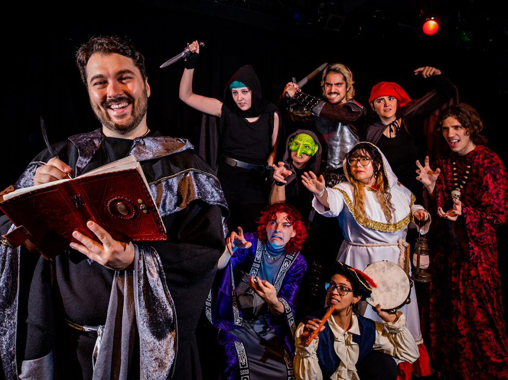 Un grupo de artistas disfrazados posa de forma teatral con accesorios de fantasía, como dagas, máscaras e instrumentos musicales, mientras un personaje ataviado con una túnica lee un libro de hechizos.