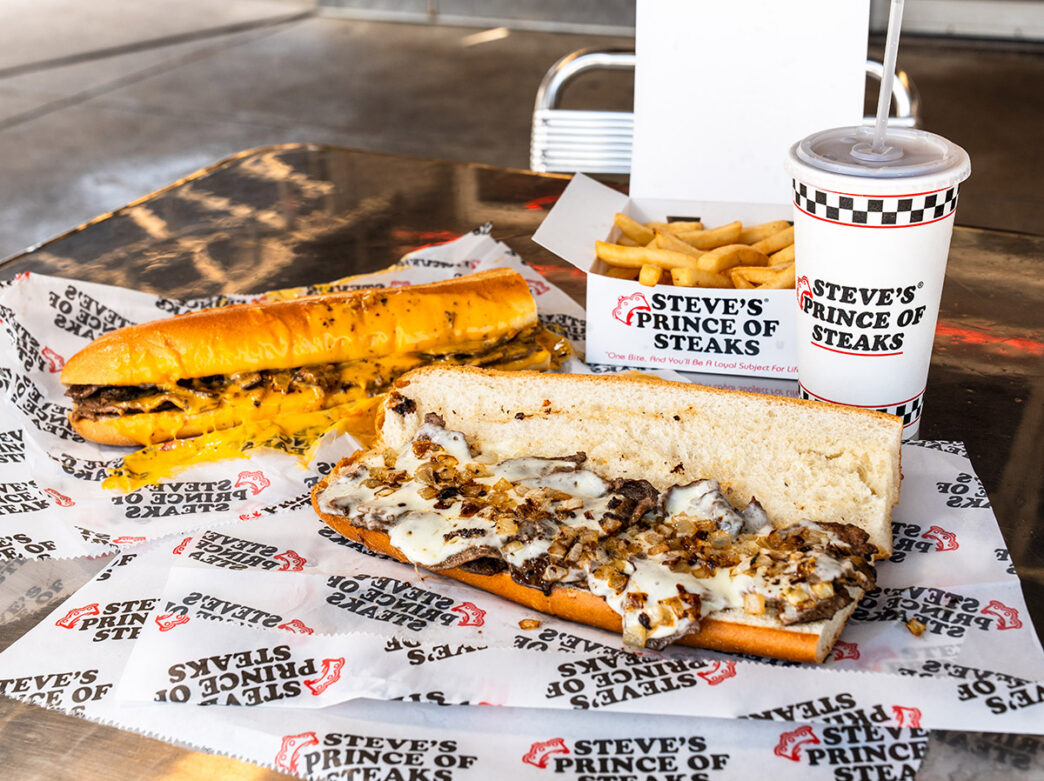 Dois sanduíches de carne com queijo estão em uma mesa de metal com batatas fritas e um refrigerante.