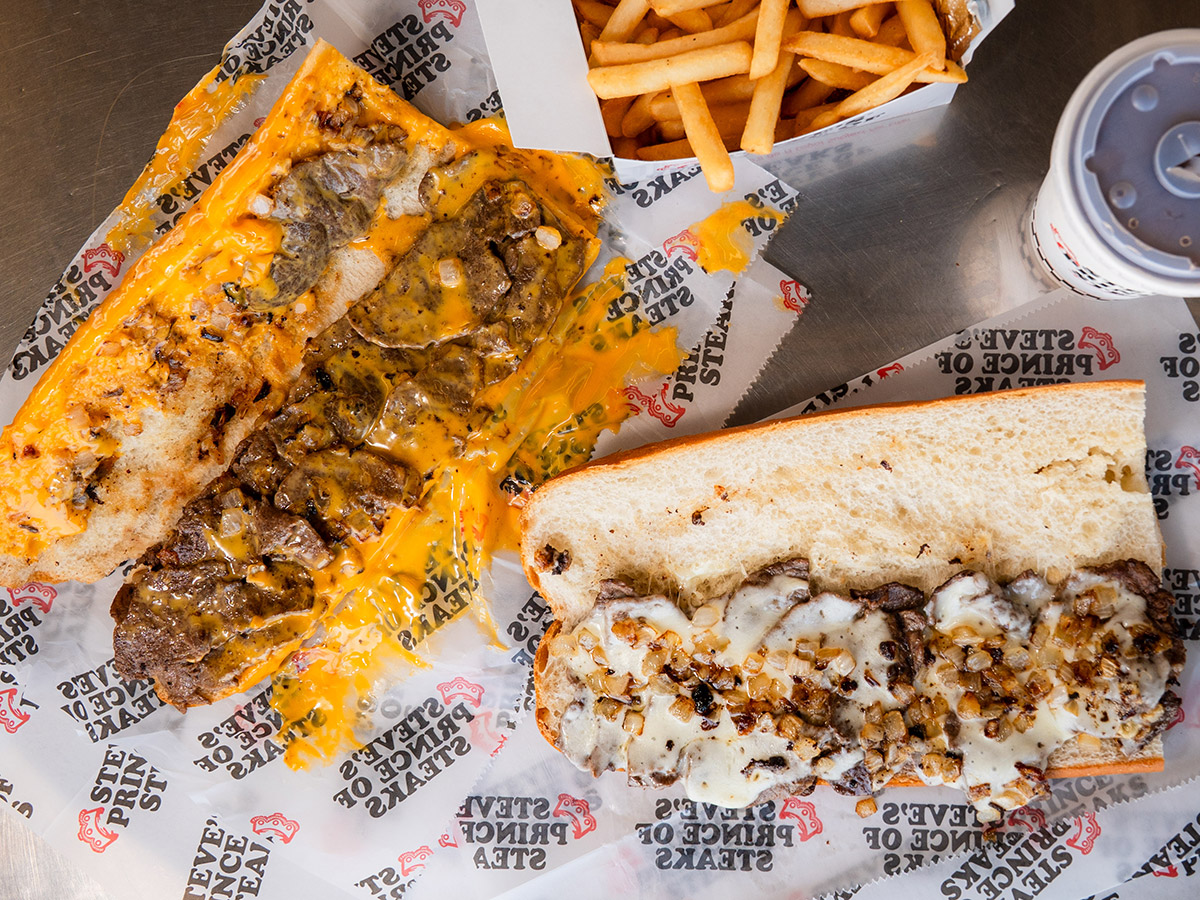 Twee cheesy steak sandwiches van Steve's Prince of Steaks, geserveerd met friet en een drankje op een metalen tafel.