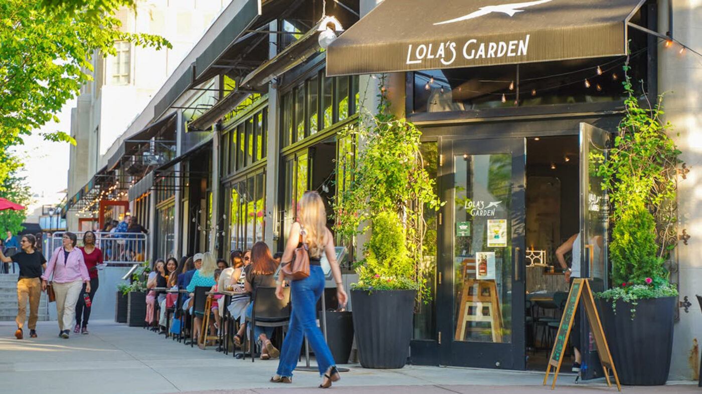 Outdoor diners sit at tables outside Lola’s Garden at Suburban Square on a sunny afternoon, while shoppers walk by.