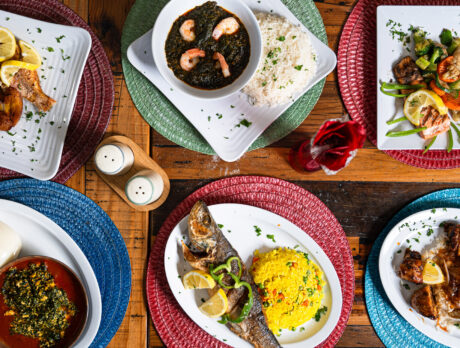 Apresentação de pratos do African Small Pot, na Filadélfia, com peixe frito inteiro com banana-da-terra, verduras refogadas com fufu, camarão ao molho escuro com arroz, frango grelhado com arroz, legumes salteados e jogos americanos de tecido de cores vivas em uma mesa de madeira.