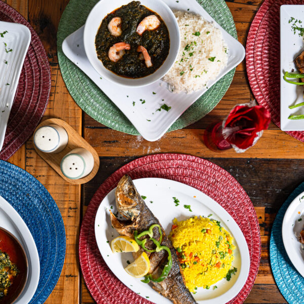 Overhead spread of dishes from African Small Pot in Philadelphia, featuring whole fried fish with plantains, stewed greens with fufu, shrimp in dark sauce with rice, grilled chicken over rice, sautéed vegetables, and brightly colored woven placemats on a wooden table.