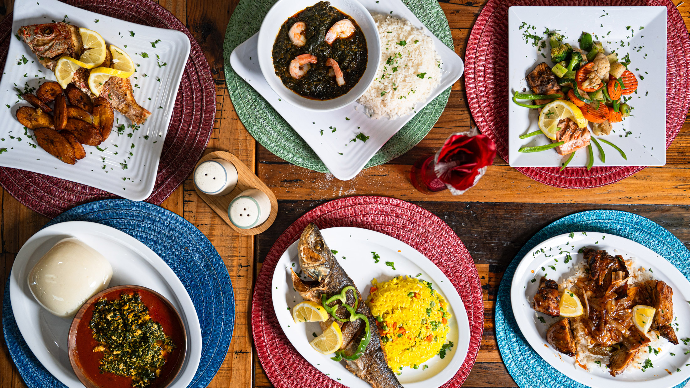 Overhead spread of dishes from African Small Pot in Philadelphia, featuring whole fried fish with plantains, stewed greens with fufu, shrimp in dark sauce with rice, grilled chicken over rice, sautéed vegetables, and brightly colored woven placemats on a wooden table.