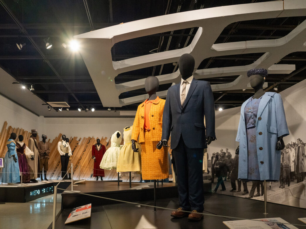 Mannequins display colorful, tailored costumes made by two-time Academy Award winner Ruth E. Carter in the Afrofuturism in Costume Design exhibit at the African American Museum in Philadelphia, with garments ranging from bright textured coats to sleek suits in a modern gallery setting.