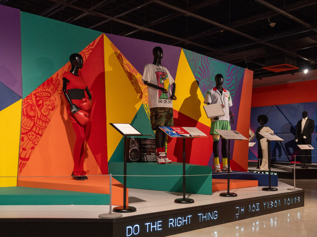 Los maniquíes exhiben los trajes de *Do the Right Thing* en la exposición «Afrofuturismo en el diseño de vestuario» del African American Museum in Philadelphia, con un fondo geométrico llamativo y colorido y carteles explicativos.