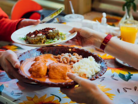 Diners pass plates of Mexican dishes at Casa Mexico in Philadelphia, including enchiladas with rice and beans and a plate of sauced chiles, set on a colorful table with drinks nearby.