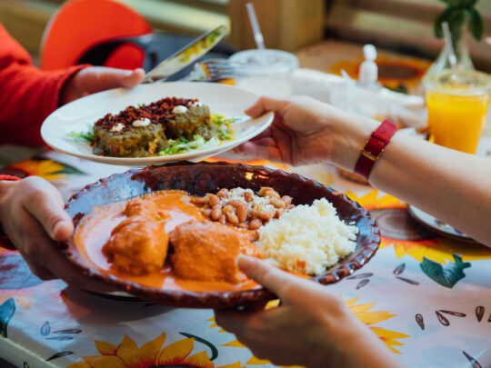 Diners pass plates of Mexican dishes at Casa Mexico in Philadelphia, including enchiladas with rice and beans and a plate of sauced chiles, set on a colorful table with drinks nearby.