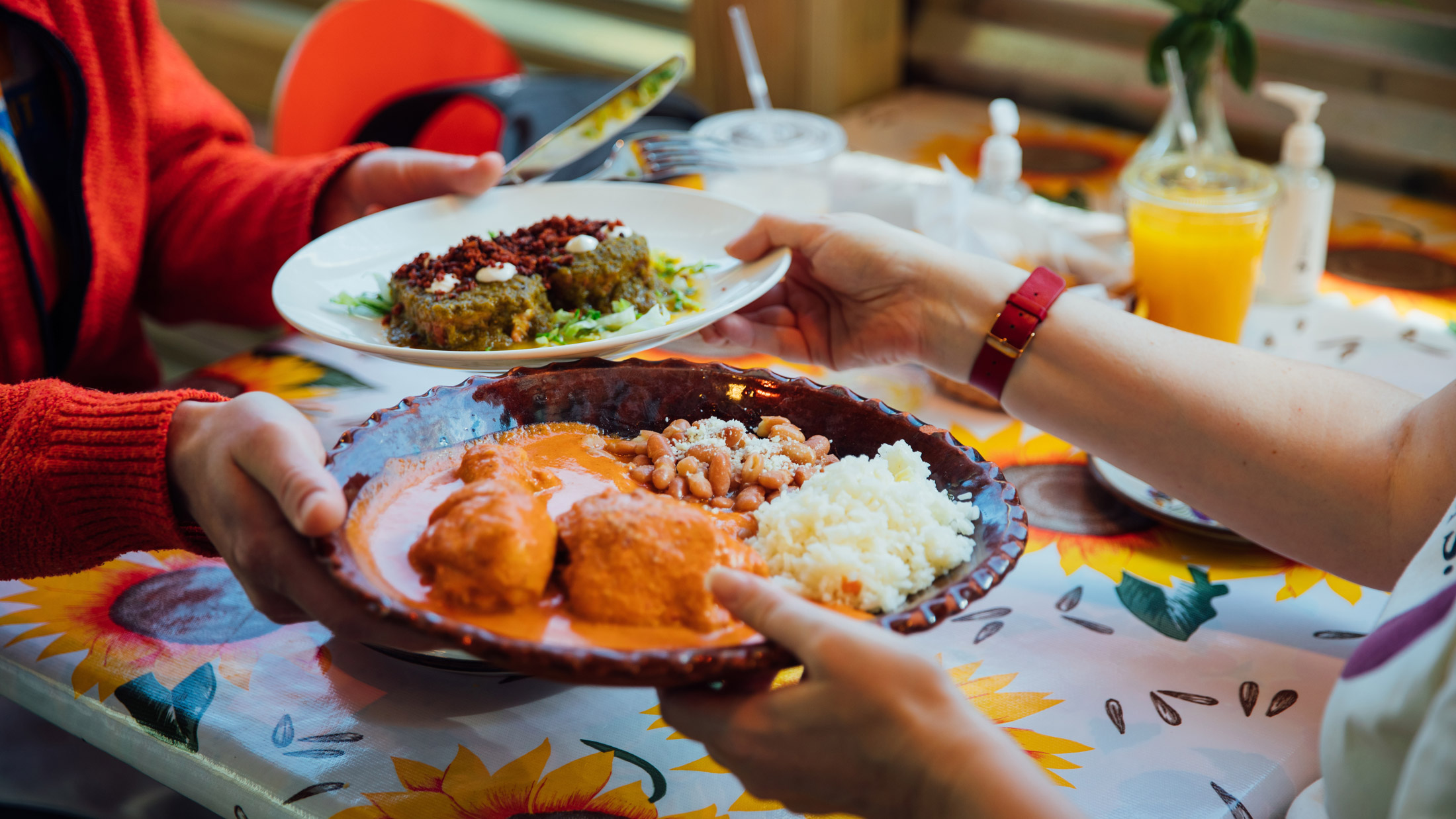 Diners pass plates of Mexican dishes at Casa Mexico in Philadelphia, including enchiladas with rice and beans and a plate of sauced chiles, set on a colorful table with drinks nearby.