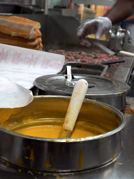 Cook preparing a cheesesteak on a griddle, with chopped beef, stacked rolls, and a pot of melted cheese sauce in the foreground.