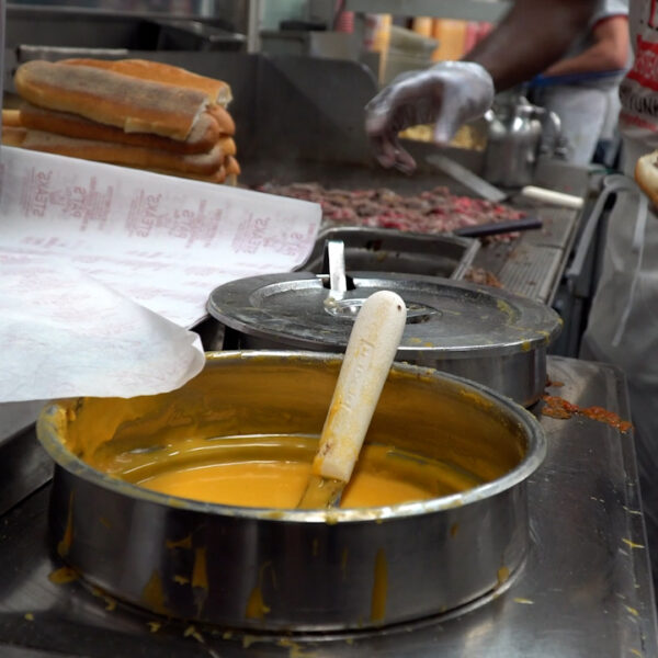 Cook preparing a cheesesteak on a griddle, with chopped beef, stacked rolls, and a pot of melted cheese sauce in the foreground.