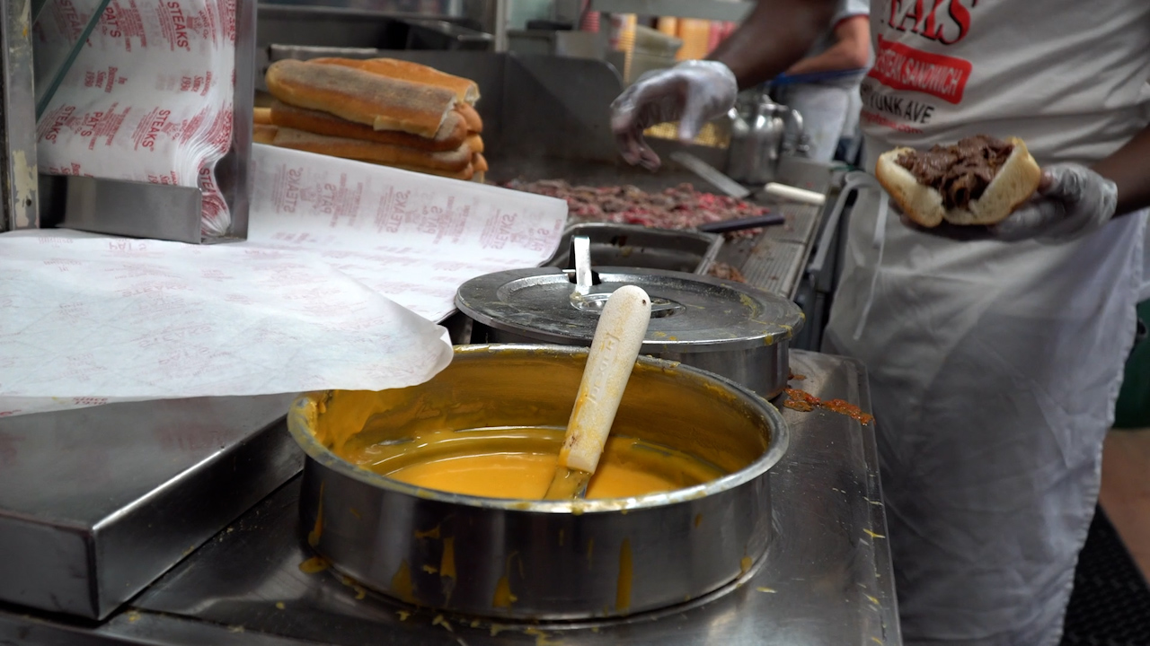 Cook preparing a cheesesteak on a griddle, with chopped beef, stacked rolls, and a pot of melted cheese sauce in the foreground.