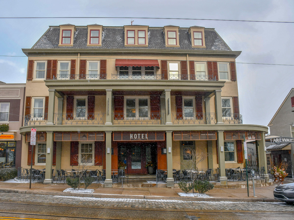The exterior of the Chestnut Hill Hotel in Philadelphia, a three-story building with shuttered windows, balconies, and a covered porch, sits along a wet street on a snowy day.