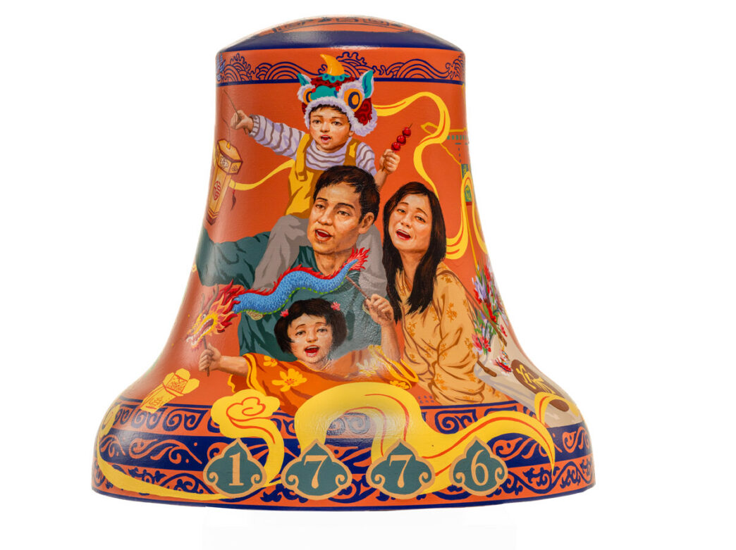 Colorfully painted bell sculpture from Philadelphia250’s Bells Across PA, featuring a family celebrating Lunar New Year. A child wearing a dragon dance headpiece sits on an adult’s shoulders, while another child holds a small dragon figure. The scene is surrounded by bright red and gold tones, stylized clouds, lanterns, and decorative patterns inspired by Chinese culture, with the numbers “1 7 7 6” displayed along the base.