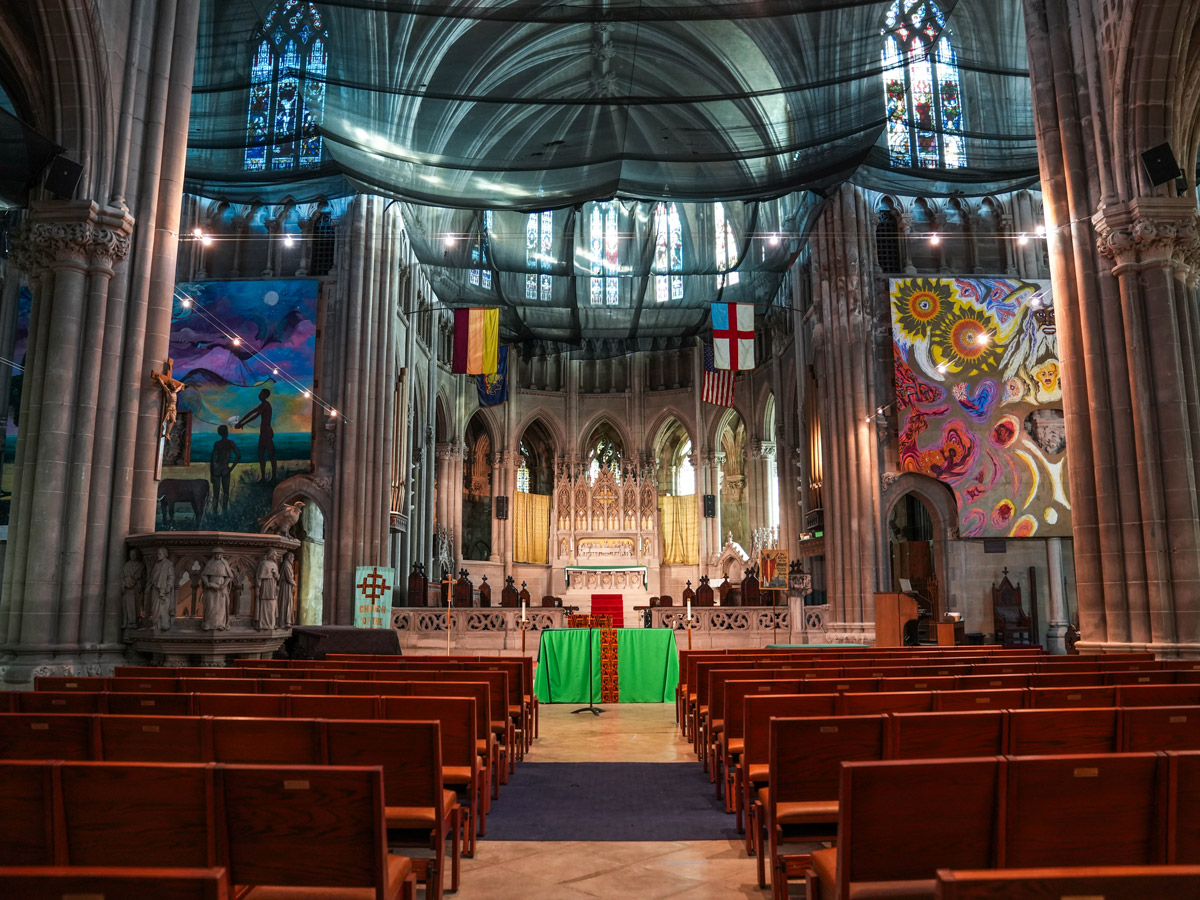 Interior of the George W. South Memorial Church of the Advocate in North Philadelphia, showing rows of wooden pews leading to an ornate altar beneath stained-glass windows and large colorful murals.