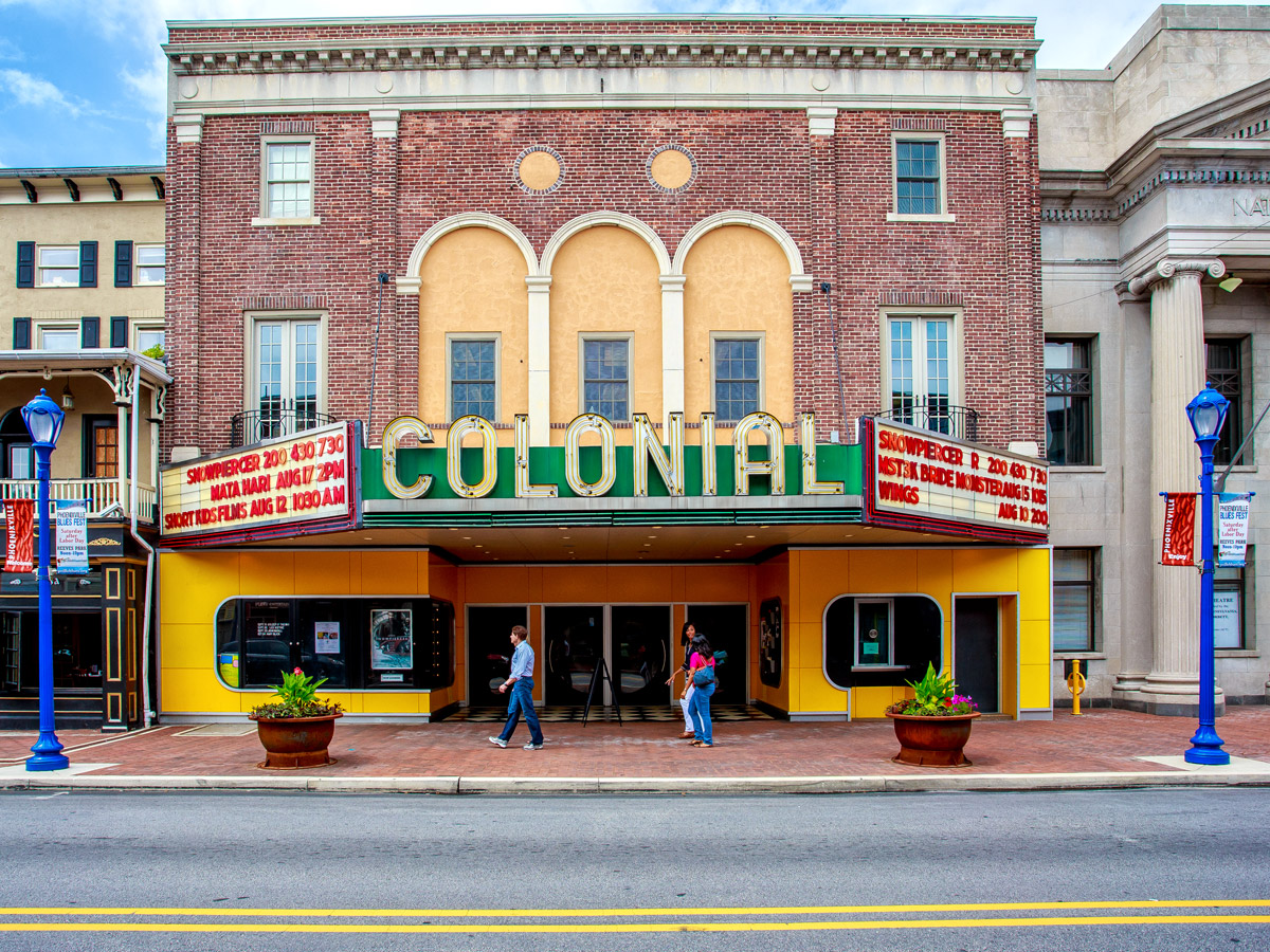 Front view of the Colonial Theatre in Phoenixville, a historic red-brick building with a bright yellow entrance and a large green-and-gold “COLONIAL” marquee, as three people walk along the sidewalk in front.
