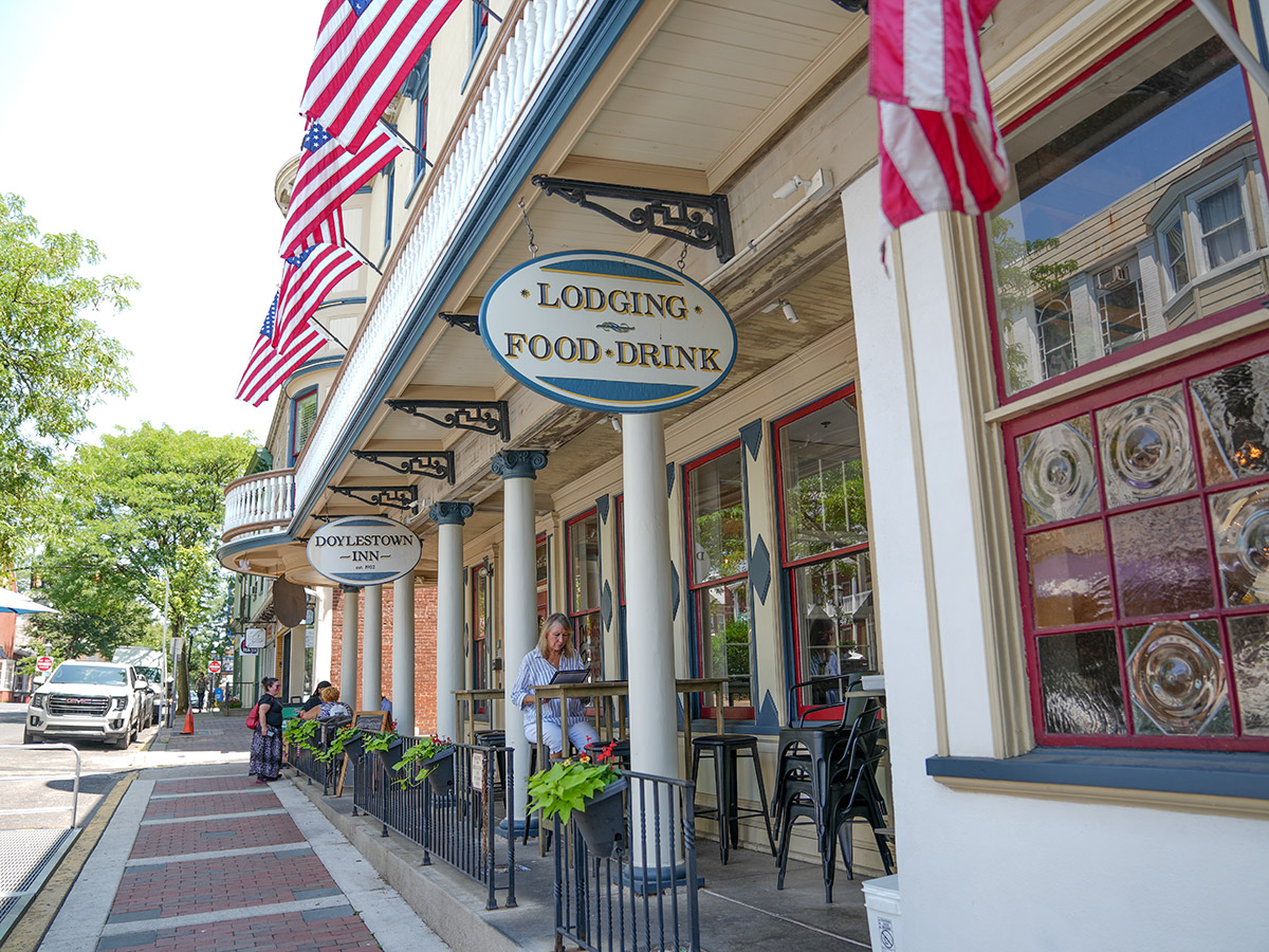 The historic Doylestown Inn features a covered sidewalk patio with outdoor seating, hanging signs and American flags lining the façade as visitors dine and stroll along the tree-lined street.