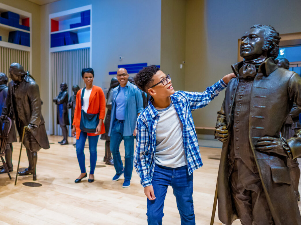 Un niño sonríe mientras interactúa con una estatua de bronce en el interior del National Constitution Center de Filadelfia, mientras dos adultos se encuentran detrás de él entre una galería de figuras históricas de tamaño natural.
