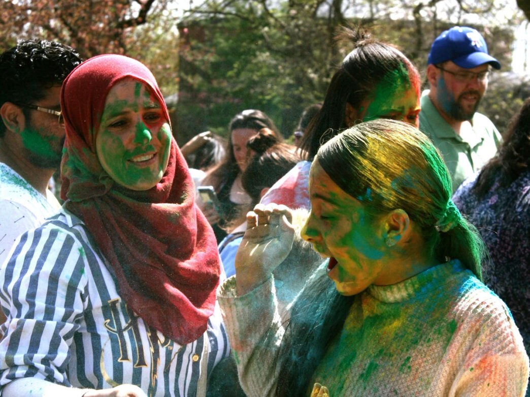 People smile and toss colorful powder during the Festival of Colors at the Philadelphia Zoo, with bright pigments covering their faces and clothing as a lively crowd gathers outdoors.