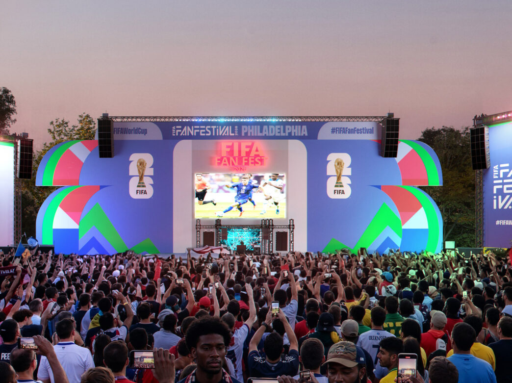 Large crowd gathered in front of a colorful stage at the FIFA World Cup 2026 Fan Festival in Philadelphia, with fans holding up phones and flags as a soccer match plays on a giant central screen.