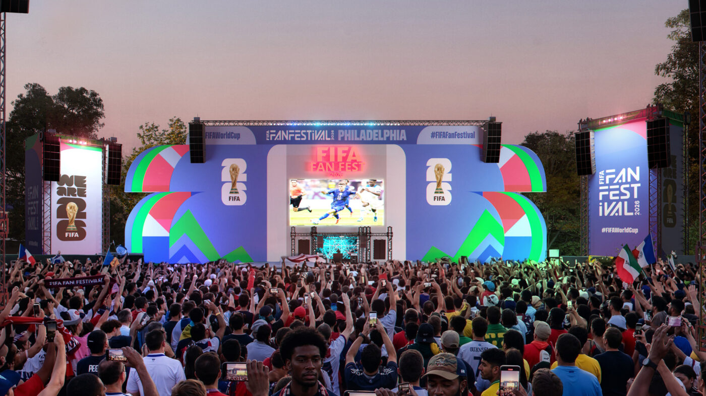 Grande multidão reunida em frente a um palco colorido no Fan Festival da Copa do Mundo da FIFA 2026 na Filadélfia, com torcedores segurando telefones e bandeiras enquanto uma partida de futebol é exibida em um telão gigante.
