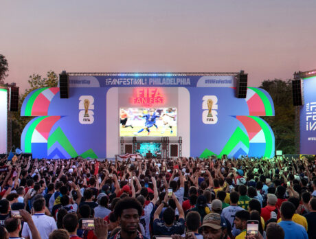 Large crowd gathered in front of a colorful stage at the FIFA World Cup 2026 Fan Festival in Philadelphia, with fans holding up phones and flags as a soccer match plays on a giant central screen.
