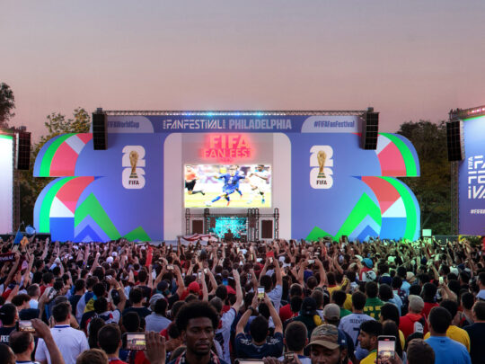 Grande multidão reunida em frente a um palco colorido no Fan Festival da Copa do Mundo da FIFA 2026 na Filadélfia, com torcedores segurando telefones e bandeiras enquanto uma partida de futebol é exibida em um telão gigante.