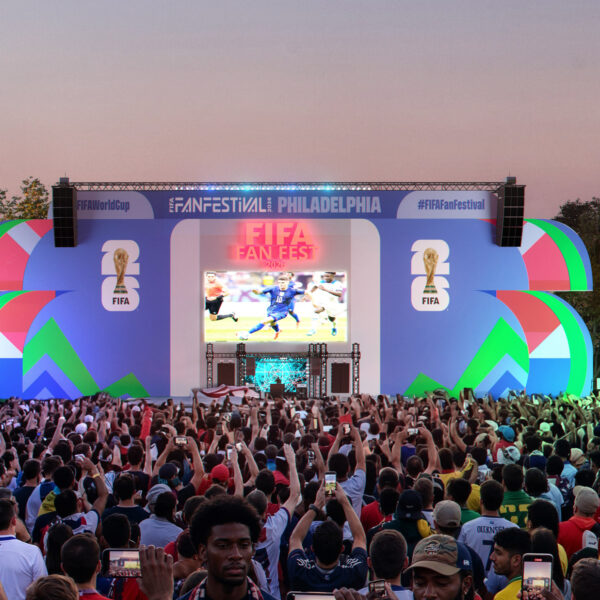 Large crowd gathered in front of a colorful stage at the FIFA World Cup 2026 Fan Festival in Philadelphia, with fans holding up phones and flags as a soccer match plays on a giant central screen.
