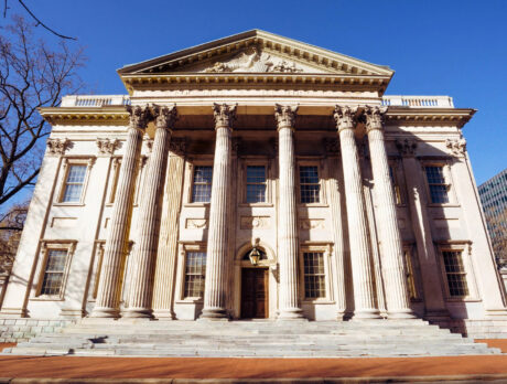 The grand front of the First Bank of the United States shows its stately row of columns and symmetrical stone architecture under a bright blue sky.