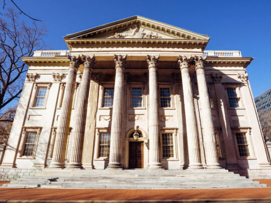 The grand front of the First Bank of the United States shows its stately row of columns and symmetrical stone architecture under a bright blue sky.