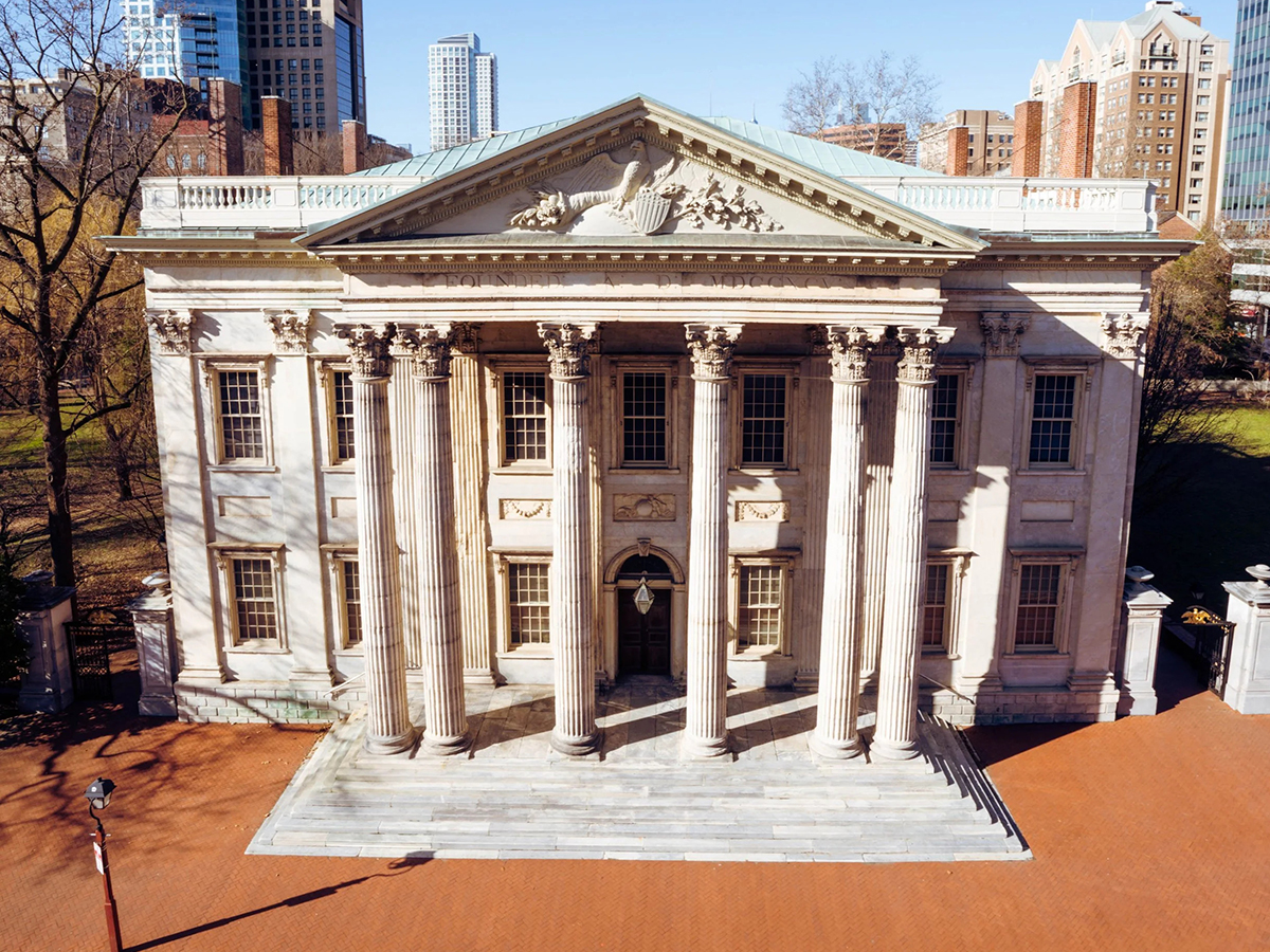The grand front of the First Bank of the United States shows its stately row of columns and symmetrical stone architecture under a bright blue sky.