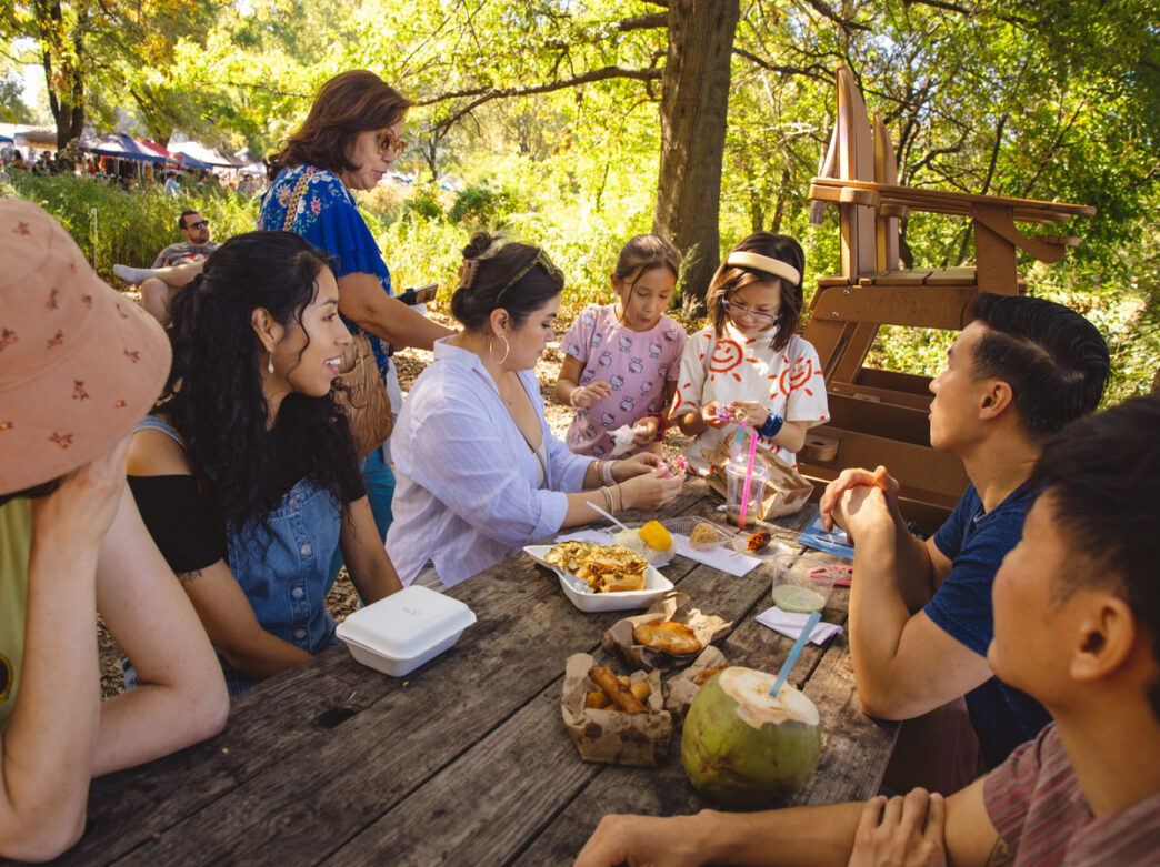 A group of adults and children gather around a picnic table at the Southeast Asian Market in FDR Park in Philadelphia, sharing food under leafy trees with vendor tents in the background.