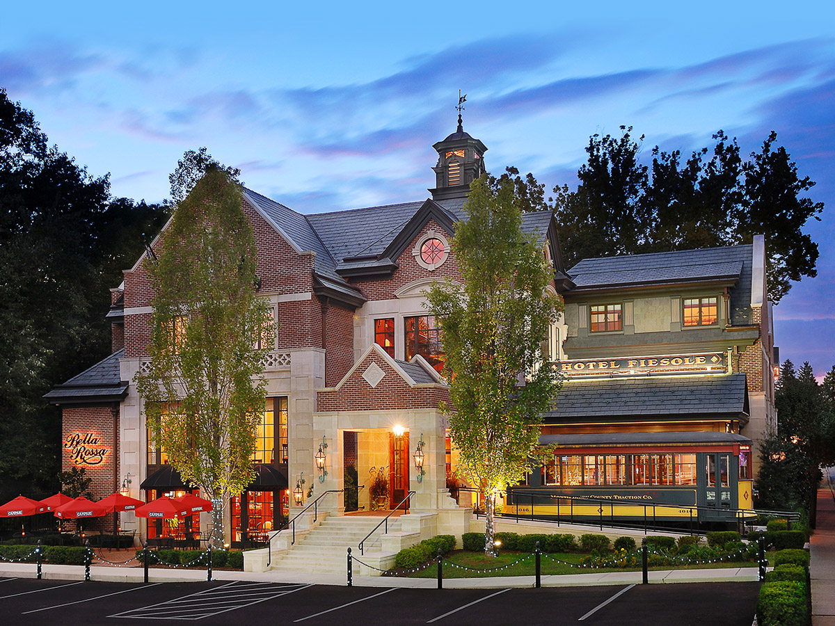 Evening view of Hotel Fiesole in Skippack, Pennsylvania, a warmly lit brick building with outdoor dining umbrellas, trees, and a vintage trolley along the side.