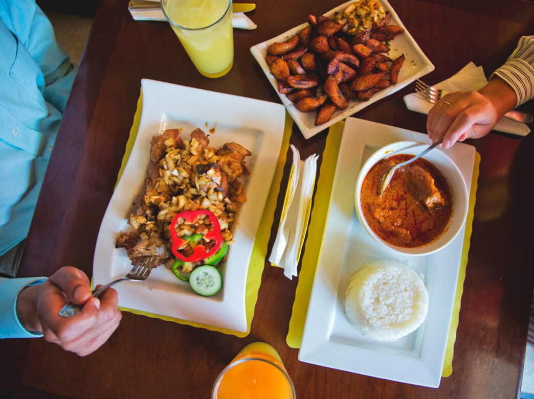 Overhead view of a table at Kilimandjaro Restaurant in Philadelphia with two diners enjoying West African dishes, including spiced meat with vegetables, a rich curry served with white rice, fried plantains, and colorful fruit drinks.