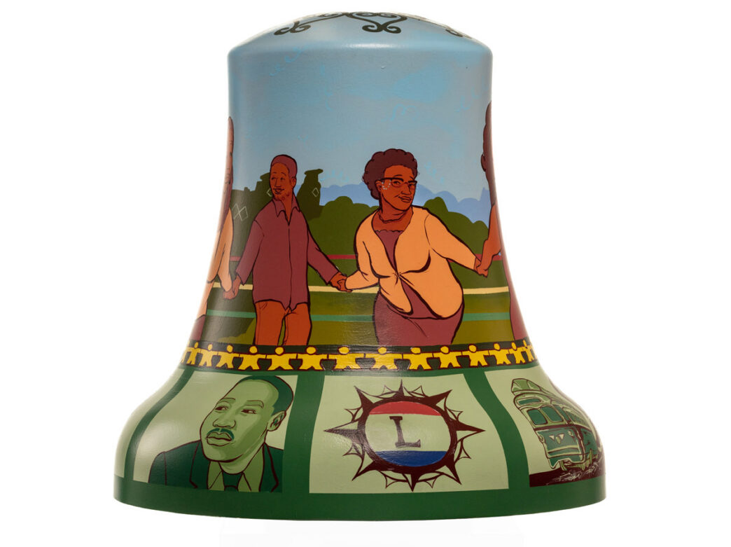Bell sculpture titled “Lancaster Living Legacy” from Philadelphia250’s Bells Across PA, featuring illustrated figures holding hands in a park-like setting against a blue sky. The lower section includes portraits and symbols tied to West Philadelphia history, including an image of Martin Luther King Jr., a trolley, and a circular emblem with an “L,” highlighting community, civil rights, and neighborhood heritage.