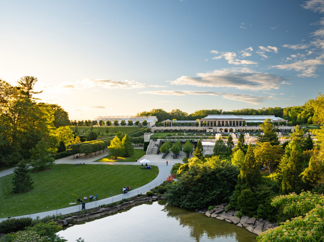 A sweeping view from Chimes Tower overlooks Longwood Gardens, with manicured lawns, winding paths, fountains, and conservatory buildings bathed in warm evening light.