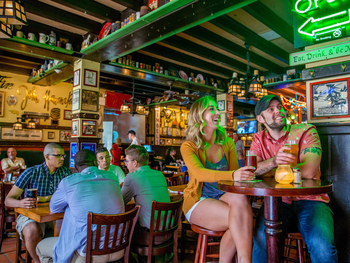 Two people sit at a high-top table drinking beers and smiling inside the lively, historic interior of McGillin’s Old Ale House in Philadelphia, with neon signs, vintage memorabilia, and other patrons socializing in the background.