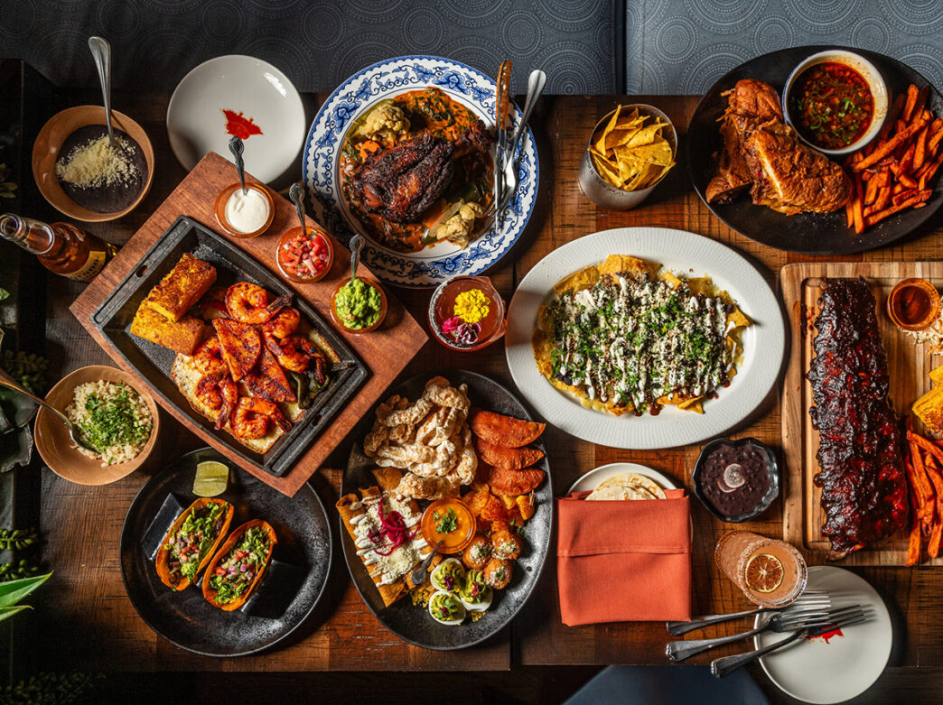 A colorful spread of Mexican dishes including tacos, grilled shrimp, nachos and ribs, fill a table alongside chips, sauces and cocktails.