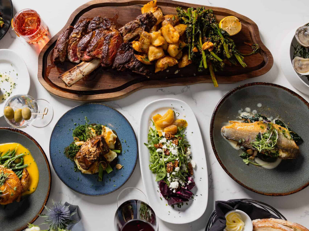 An elegant spread of plated dishes, including steak, fish and vegetables, surrounds a large tomahawk steak board at Maddy Rose at the Landing.