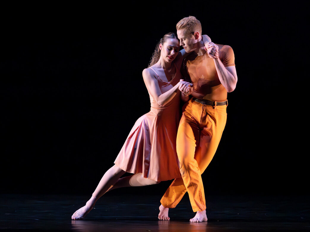 Dos bailarines interpretan un dúo íntimo sobre un escenario a oscuras, con sus cuerpos inclinados el uno hacia el otro en un movimiento elegante y sincronizado.