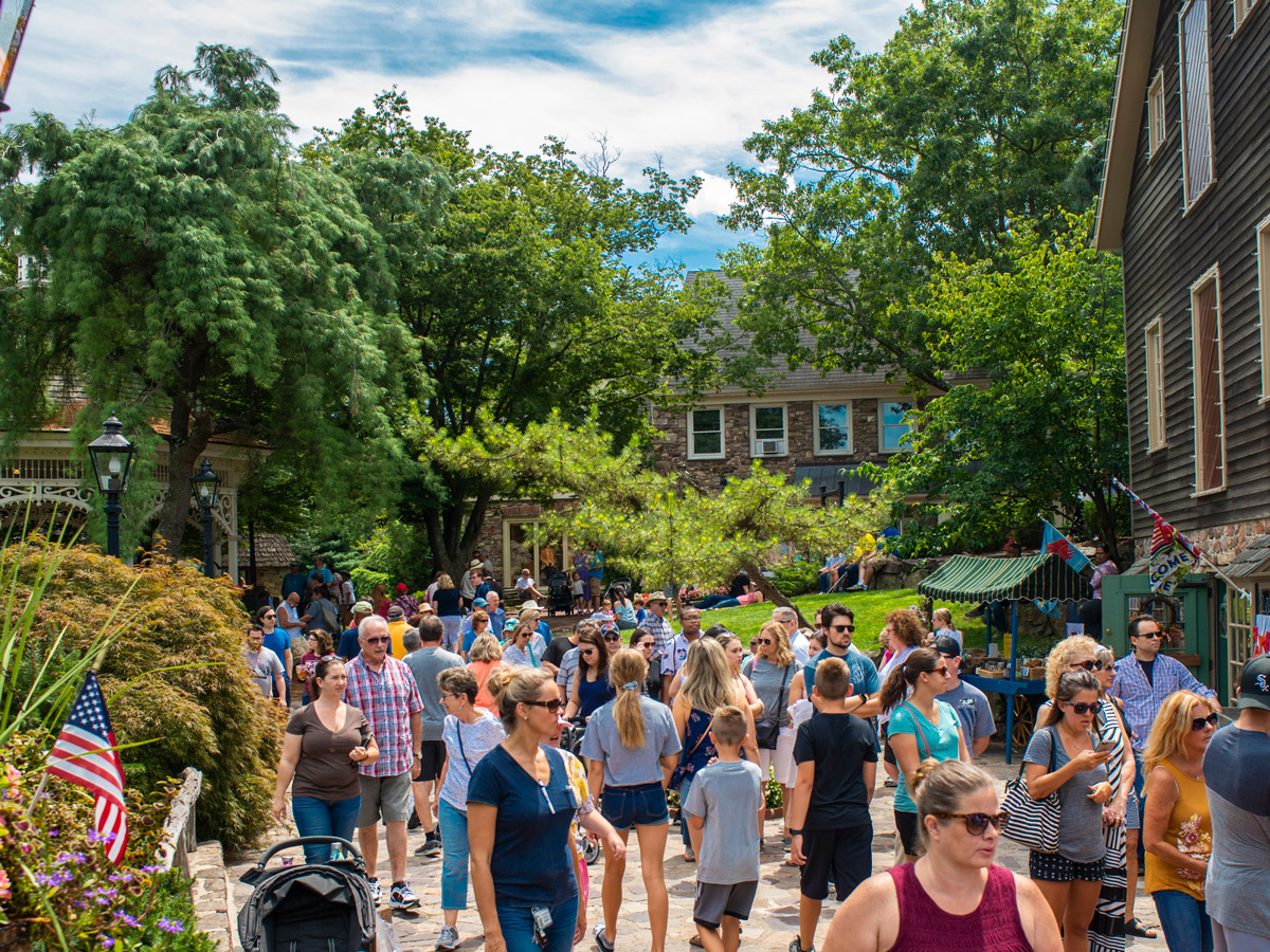 Crowds of visitors stroll along a tree-lined pathway at Peddler’s Village in Pennsylvania, passing shops, garden landscaping and outdoor vendors on a sunny day.