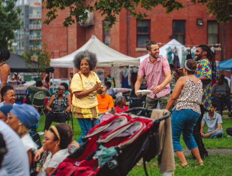 People dance and gather on a grassy lot during the Lancaster Avenue Jazz Fest in West Philadelphia, with vendor tents, clothing racks and a brick building in the background.