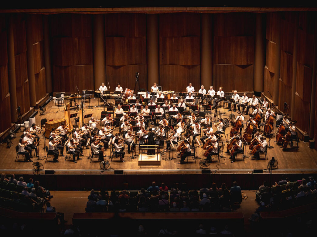 Dos bailarines interpretan un dúo íntimo sobre un escenario a oscuras, con sus cuerpos inclinados el uno hacia el otro en un movimiento elegante y sincronizado.