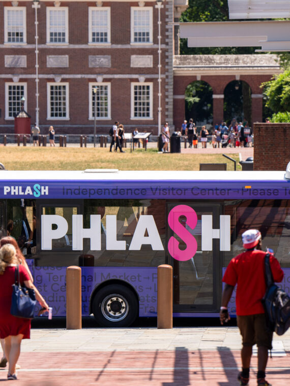 Philadelphia’s PHLASH bus is parked near Independence Mall as visitors walk along the sidewalk, with historic buildings and open green space visible in the background.